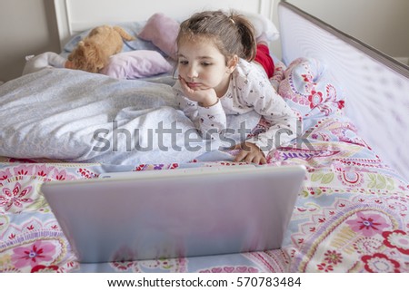 Little girl lying in bed and watching cartoons with a laptop in his bedroom. She looks entertained