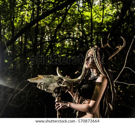 portrait of woman with dreadlocks hair hold in hand staff with cow skull with horn against wild forest trees Young girl look at God or devil. Woman shaman in ritual garment stand from fur and leather