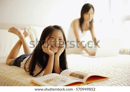 Mother using laptop, daughter lying down with book open in front of her