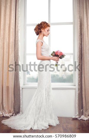 Wedding. Beautiful bride indoors with bouquet of flowers against big window in full lenght
