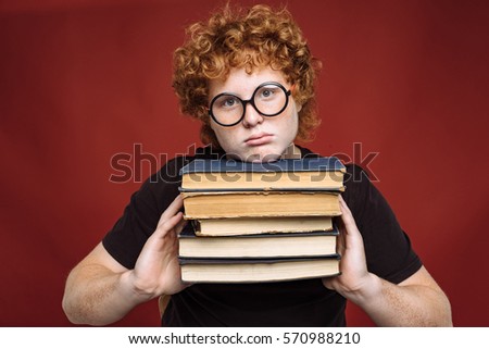 Funny shot of Nerdy curly red-haired freckled teenager holding books on red background