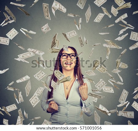 Portrait happy woman in glasses exults pumping fists celebrates success under money rain falling down dollar bills banknotes isolated on gray wall background 