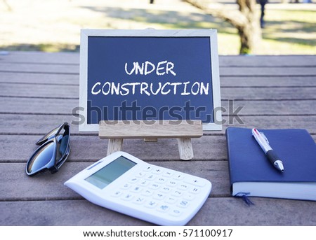 Blackboard with calculator, notebook, ball pen and sunglasses on wooden table. UNDER CONSTRUCTION                           