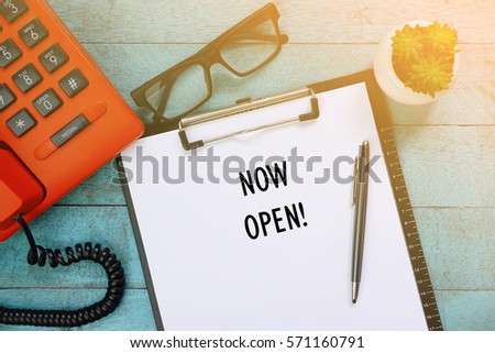 Top view of clipboard and white sheet written with NOW OPEN! on blue wooden background. Business Concept.