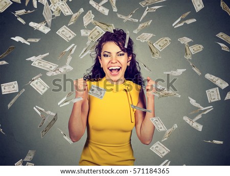 Portrait happy woman pumping fists ecstatic celebrates success under a money rain falling down dollar bills banknotes isolated on gray wall background with copy space  