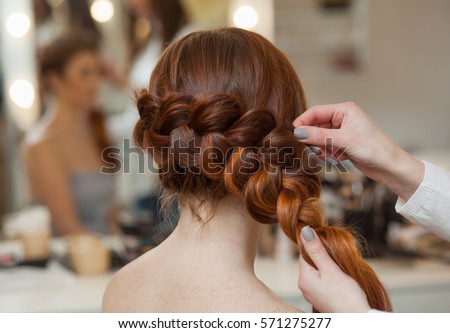 Beautiful, with long, red-haired hairy girl, hairdresser weaves a French braid, close-up in a beauty salon. Professional hair care and creating hairstyles. Royalty-Free Stock Photo #571275277