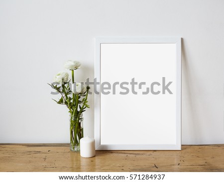 Empty frame mockup for design presentation, bouquet of flowers ranunculus and white candle on a white wall background and wooden table. Romantic minimalism design.