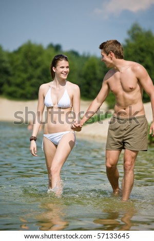Happy couple in swimwear walk in lake, coastline in background