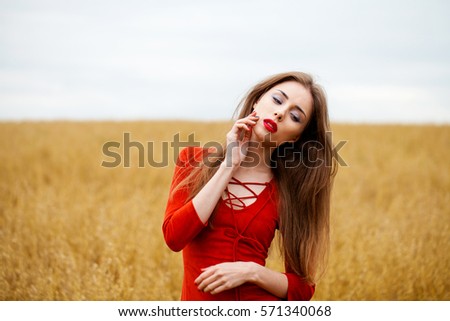Portrait of a young brunette woman in red dress on a background of golden oats field, summer outdoors