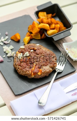 Grilled minced steak of beef with onions and fried potatoes on a black plate. Pleskavitsa - Serbian dish.