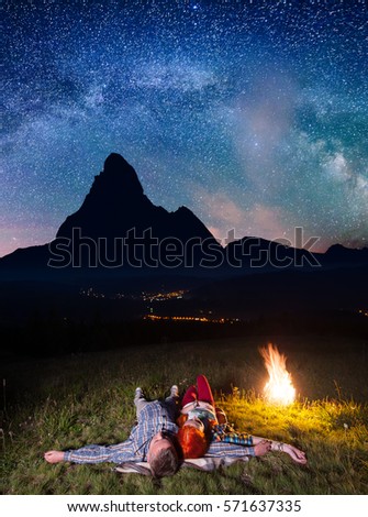 Happy couple hikers admiring the bright stars and lying near the fire at night. In the background silhouette of the mountains and luminous village in the valley. Long exposure