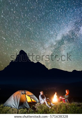 Night tent camping. Tourist family - man and woman sitting by bonfire under incredibly beautiful starry sky. In the background silhouette of the high mountains and luminous village in the valley
