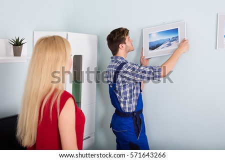 Young Woman Showing Male Carpenter For Hanging Picture Frame On Wall At Home
