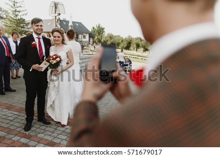 man taking photo on phone of luxury happy wedding couple of newlyweds after ceremony at church. stylish groom and bride holding certificate register. funny moment.