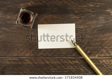 An overhead photo of a blank business card on a dark wooden background texture with a golden ink pen and ink bottle. A mockup or a banner with copy space