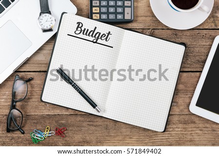 Budget written in note pad, office desk with electronic devices, computer and paper, wood table from above, concept image for blog title or header image.