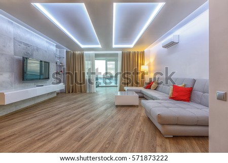 Contemporary spacious living room with hardwood floor, white ceiling, golden curtains, grey innovative coach, red decorative pillows. Sophisticated lightning. Royalty-Free Stock Photo #571873222