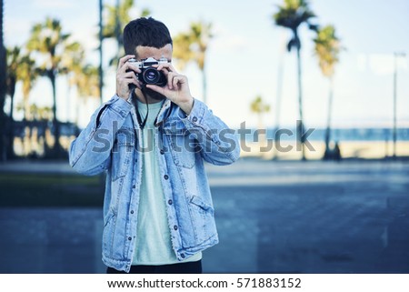 Stylishly dressed male photographer hipster taking picture of his vacation abroad  fascinated by city scape using vintage camera to convey atmosphere of coastline with palm trees strolling on streets
