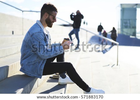 Young male hipster traveler dressed in street wear  enjoying trip during day off taking selfie and posting picture in social networks to share impressions with friends using smartphone and 4G internet