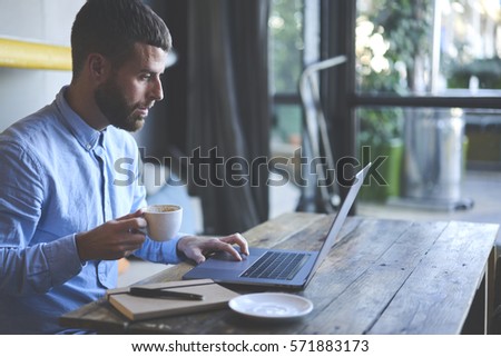 Young skilled male freelancer starting working day early in morning drinking coffee to wake up making new design for website with new advertising content using modern laptop computer and wifi