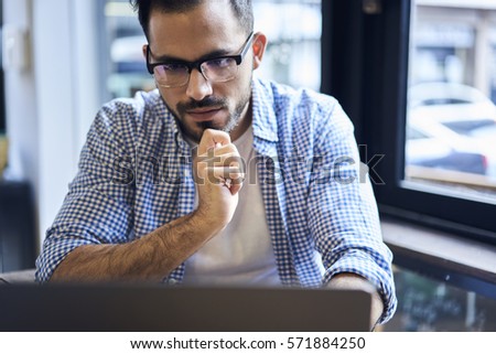 Close up portrait of mindful male blogger in eyewear watching streaming video of workshop to write feedback article on personal web page concentrated on work using laptop and wireless internet