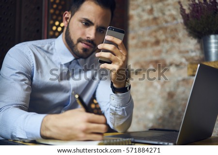 Young talented bearded male journalist noting address of meeting with successful entrepreneur to write interview while sitting in coworking space using modern technologies and wireless connection