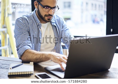 Concentrated male IT programmer brainstorming while coding new software for digital devices sending his fork for testing using modern laptop computer and wireless connection to internet in office