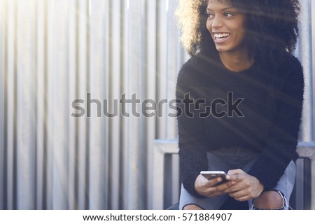 Cheerful attractive afro American female travel blogger feeling excited after reading feedback from followers while sitting outdoors using free wireless connection to internet and modern smartphone