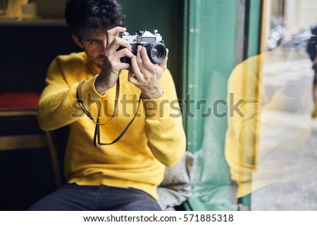 Male hipster tourist dressed in trendy clothing taking picture on his professional vintage camera while sitting in urban coffee shop near the window.Male photographer making photos for his travel blog
