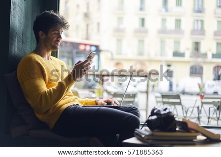 Creative male photographer working in modern cafe choosing best images for publishing in blog texting messages with executive using online chat on smartphone connected to free wireless internet Royalty-Free Stock Photo #571885363