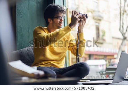 Young male fashion blogger and profesional photographer getting inspire with trendy looks of people outside on street is taking picture on his vintage camera while sitting in cozy cafe near window