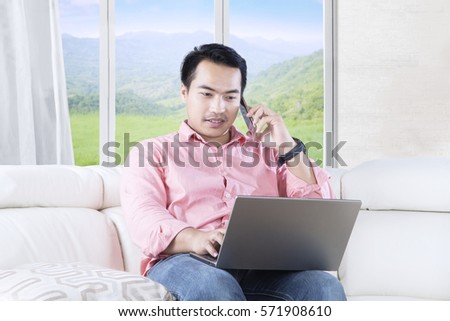 Image of a modern man using a laptop while sitting in the living room and talking on the mobile phone