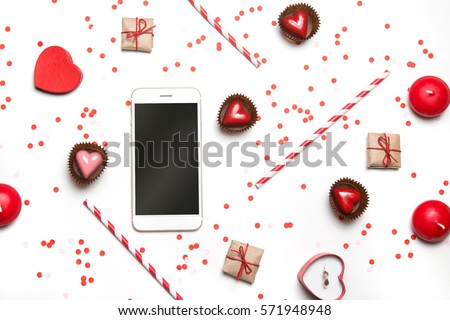 Styled photo of St. Valentine's Day flatlay top view isolated on white. Red heart sweets, gifts in craft paper, proposal ring in box, smartphone mockup, candles and confetti. 
