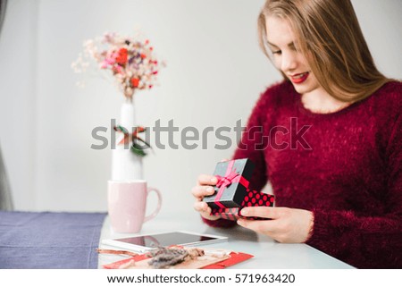 Young pretty smiling blonde girl with maroon lips and sweater sitting at the table and opening a gift. Card, flowers, tablet on background. Valentine's Day, Christmas, Birthday, Holidays concept.