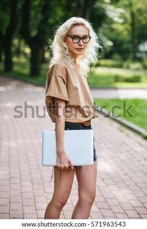 Blonde pretty woman in glasses standing with her laptop in park in summer. Portrait of a smiling student on break outdoors. Lifestyle photo