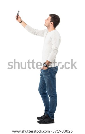 Side view of young handsome man wearing light gray shirt taking selfie with mobile phone. Full body length portrait isolated over white studio background. 
