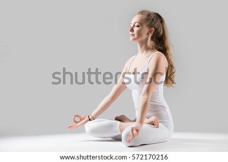 Young attractive woman practicing yoga, sitting in Padmasana exercise, Lotus pose on meditation session, working out wearing sportswear white tank top, full length, isolated, grey studio, closed eyes  Royalty-Free Stock Photo #572170216