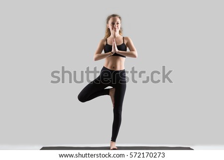 Young attractive woman practicing yoga, standing in Vrksasana exercise, Tree pose, working out, wearing sportswear, black sports bra, pants, indoor full length, isolated against grey studio background