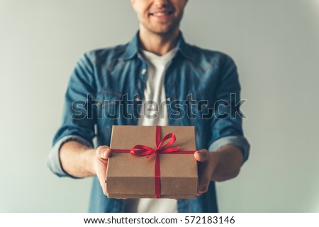 Cropped image of handsome romantic guy smiling while holding a present for his couple, on gray background