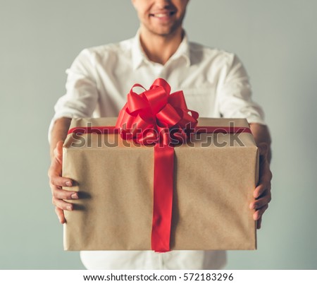 Cropped image of handsome romantic guy smiling while holding a big gift box for his couple, on gray background