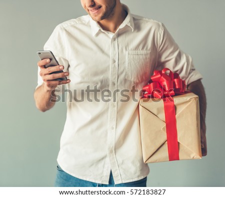 Cropped image of handsome romantic guy using a smartphone and smiling while holding a big gift box for his couple, on gray background