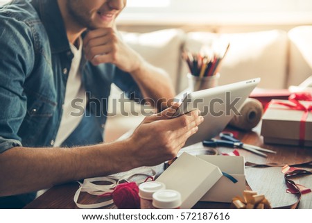 Cropped image of handsome romantic guy using a digital tablet and smiling while making present for his couple