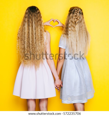 Love and friendship forever. Two girls girlfriends standing back and show making heart sign. Indoor. On yellow background.