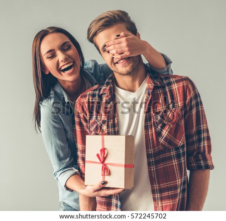 Beautiful girl is covering her boyfriend eyes and giving him a present, on gray background Royalty-Free Stock Photo #572245702