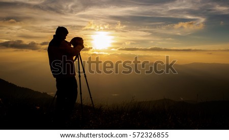 Silhouette man photographer using a tripod taking photo on sunset mountain peak.