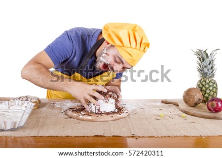 Picture of cheerful male chef in yellow uniform eating whole cake isolated on white background