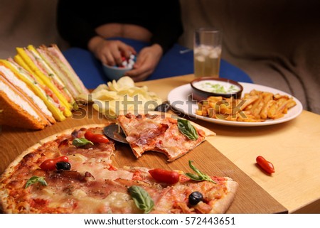 Fat man sitting on a sofa eating candy at a table with pizza, sandwiches, chips and fries. is unhealthy lifestyle.  junk food.