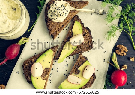 Sandwiches with avocado, radish, arugula, cheese and nuts on a rye bread with sunflower seeds for healthy breakfast on dark wooden background. Vegetarian food. Top view.