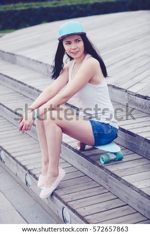 Happy young skater girl sit on skateboard deck in park outdoor.Good summer weather,attractive female model skateboarder.Vertical shot of friendly smiling chick in jeans shorts and white tank top shirt