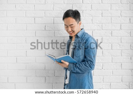 Young Asian man with book standing near brick wall